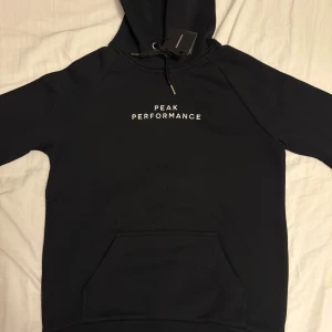 Svart hoodie från Peak Performance - Svart hoodie från Peak Performance med vit text på bröstet. Klassisk modell med huva, dragsko och stor magficka. Tillverkad i mjuk bomullsmix, perfekt för chill eller streetwear. Snygg och enkel design som funkar året runt. Aldrig tidigare använd. Ny pris ca 1200