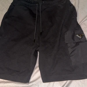 Prada shorts - Svarta shorts från Prada med cool ficka på sidan och klassisk logga. De har snörning i midjan och är tillverkade i mjuk bomull med detaljer i syntet. Perfekta för dig som gillar stilrena och exklusiva plagg med streetkänsla.
