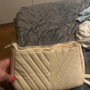 Beige quiltad axelväska med dragkedja - Säljer en stilren beige axelväska med quiltad design och dragkedja upptill. Väskan har en enkel och snygg look som passar till många outfits. Perfekt storlek för det viktigaste och lätt att bära över axeln.