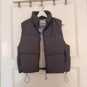 Grå väst från Bershka XS - Grå väst från Bershka i storlek XS med hög krage och dragkedja framtill. Västen har justerbara snören nertill och ett tryck på insidan med texten 'LOVE OVERLOAD YOU JUST GOT LUCKY' i blått. Perfekt för lager-på-lager-stil.