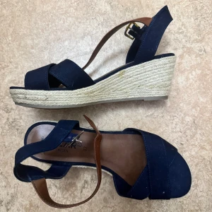 Marinblå kilklack sandaler med rem - Snygga marinblå sandaler med kilklack i rep och bruna remmar med spänne. Har aldrig använt dessa vilket man kan se, storlek 37 och om jag inte har fel så är de från dinsko. 
