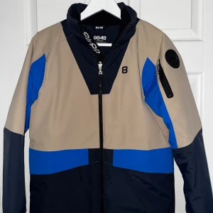 8848 beige och blå jacka - Snygg jacka från 8848 i beige, blått och mörkblått. Jackan har dragkedja framtill, hög krage och en ficka med dragkedja på ärmen. Sportig design med färgblock och logga på bröstet och ärmen.