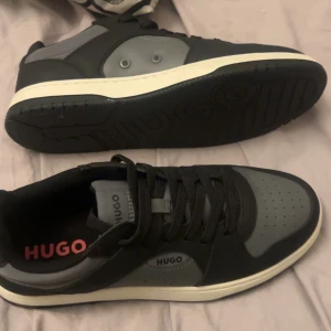 Svarta och grå sneakers från Hugo - Snygga sneakers från Hugo i svart och grått med vita sulor. Skorna har klassisk rund tå, svarta snören och diskret Hugo-logga på sidan och plösen. Ovandelen är i syntetmaterial och designen är stilren med perforerade detaljer framtill.