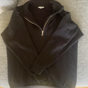 Svart half zip sweatshirt Green Day - Svart sweatshirt från Green Day med halv dragkedja och hög krage. Tröjan har en clean look och är tillverkad i mjuk bomull, perfekt för lager-på-lager. Passformen är normal och ärmarna är långa. Enkel att matcha med jeans eller joggers.