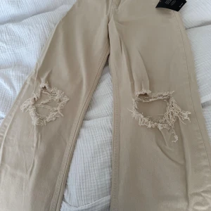 Beige ripped jeans från nakd - Beiga jeans från Romee Strijd x NA-KD i ekologisk bomull. Modellen har raka/vida ben, hög midja och stora slitningar vid knäna för en cool, avslappnad look. Färgen är cream och materialet känns mjukt men ändå stadigt. 