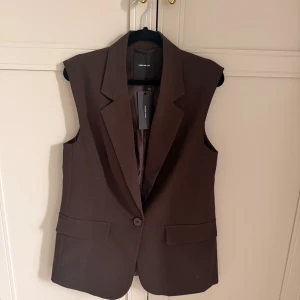 Brun kostymväst från Vero Moda - Stilren brun väst från Vero Moda med klassisk kavajkrage och en knapp framtill. Västen har två fickor med lock och är figursydd för en snygg siluett. 