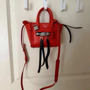 Röd handväska Zadig & Voltaire - Cool röd handväska från Zadig & Voltaire i skinn med silverfärgade metalldetaljer, dubbla dragkedjor och hängande svarta remmar. Väskan har både handtag och justerbar axelrem, samt ett snyggt hänglås framtill. Perfekt för dig som gillar edgy design.