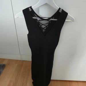 Svart klänning med snörning - Svart ärmlös klänning från Vinceotto med tight passform. Köpt i Spanien. Klänningen har v-ringning med nätmaterial och snörningsdetalj baktill. Materialet är stretchigt och bekvämt.