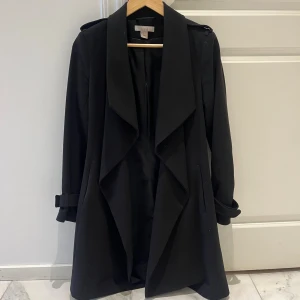 Svart kappa från H&M, strl 34 - Stilren svart kappa från H&M i storlek 34. Jackan har bred krage, snyggt fall och detaljer med guldiga öljetter vid ärmsluten. Perfekt för dig som gillar en klassisk look med modern twist.