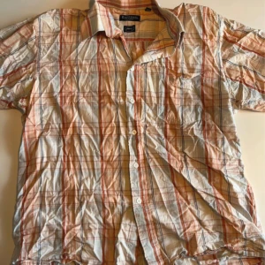 Rutig skjorta från Rappson XL - Rutig skjorta från Rappson i ljusa färger med inslag av orange, blått och beige. Klassisk krage, kort ärm och knäppning framtill. Skjortan är i bomull och har en bröstficka. Perfekt för dig som gillar en avslappnad stil.