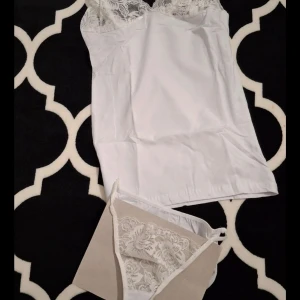Vit linnetopp med spetsdetaljer 38-40 - Snygg vit linnetopp med tunna axelband och vacker spets upptill. Toppen har en feminin v-ringning och är tillverkad i ett mjukt material som känns skönt mot huden. Perfekt att styla med jeans eller kjol för en fräsch look.