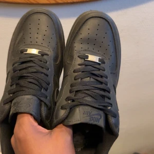 Nike Air Force 1 helsvarta sneakers - Säljer ett par klassiska Nike Air Force 1 i helsvart skinn. Låga sneakers med svart sula, svarta skosnören och diskreta detaljer i metall framtill. Perfekta för dig som gillar en clean och tidlös look. Ikonisk Nike-logga på sidan och tungan. Skorna är använda 2 gånger inga skador skicket är på 9,5/10 hör av er vid pris eller fler bilder.