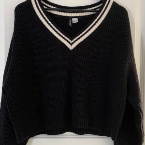 Svart stickad tröja med v-ringning Divided - Svart stickad tröja från Divided med bred v-ringning och vita ränder runt halsen. Tröjan har en loose passform, ribbade muddar och långa ärmar. Perfekt för lager-på-lager och chill vibes.