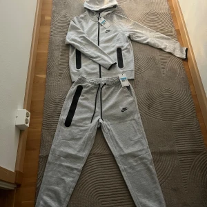 Nike tech  - Snygga grå mjukisbyxor från Nike med svarta dragkedjor på fickorna och svart snörning i midjan. Byxorna har en relaxed fit och är gjorda i mjukt bomullsmaterial. Perfekta för chill eller träning, med klassisk Nike-logga på benet.