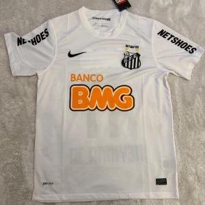 Vit Santos Neymar Jr fotbollströja Nike - Santos FC fotbollströja i vitt med Neymar Jr och nummer 11 på ryggen. Tröjan har korta ärmar, svarta tryck och orange BMG-logga. Märkt med Nike Swoosh och Dri-FIT-teknologi för extra komfort. Sponsorer på ärmar och bröst. Perfekt för fans av brasiliansk fotboll.