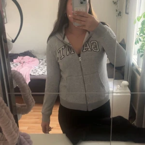 Grå hoodie från GANT med dragkedja - Grå hoodie från gang med broderad logga framtill. Tröjan har dragkedja, huva och två fickor. Tillverkad i mjuk bomullsblandning och har ribbade muddar vid ärmslut och nederkant. Inte använd på länge. Det är inte en fläck på bild tre det är bara skuggan.