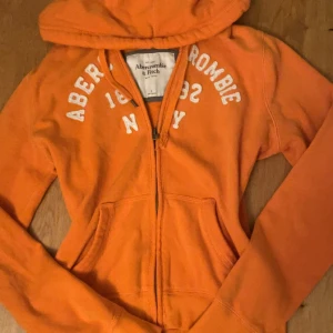 Orange hoodie från Abercrombie & Fitch - Säljer en snygg orange hoodie från Abercrombie & Fitch med vit text på bröstet. Tröjan har huva, dragkedja och två fickor framtill. Perfekt för chill dagar och har en klassisk streetstil. Långärmad och i mjuk bomull. lite lite mörkare färg i verkligheten. står att det är en L på tröjan men själv har jag M och den passar rätt bra
