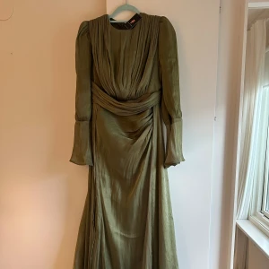 Olivgrön långklänning med vida ärmar - Olivgrön långklänning i glansigt, lätt material med draperad överdel och markerad midja. Klänningen har långa, vida ärmar och vackert fall. Perfekt för dig som vill sticka ut med en elegant och unik look.