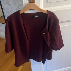 Vinröd cardigan kofta från Gina Tricot - Kort vinröd cardigan kofta från Gina Tricot med öppen front och en liten guldfärgad knappdetalj. Koftan har korta ärmar och en loose passform, perfekt för lager-på-lager. Mjuk och skön känsla i materialet.