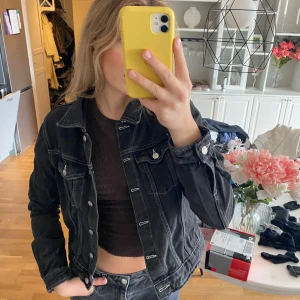 Svart jeansjacka med knappar - Snygg svart jeansjacka med kontrasterande vita sömmar och silverfärgade knappar framtill. Klassisk modell med två bröstfickor med lock och knapp, samt långärmad passform. Perfekt att slänga över en t-shirt för en chill look.