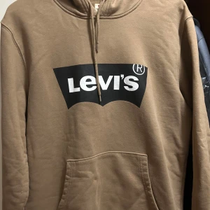 Beige hoodie från Levi's med logga - Säljer en beige hoodie från Levi's i storlek S. Tröjan har stor svart och vit Levi's-logga på bröstet, snörning vid huvan och en klassisk känguruficka framtill. Perfekt för dig som gillar streetwear och vill ha en chill look.