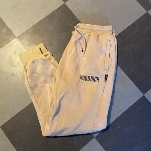 Beige mjukisbyxor från Hoodrich - Beiga mjukisbyxor från Hoodrich med svart logga på låret. Byxorna har resår och snörning i midjan samt muddar vid bensluten. Synliga tecken på användning