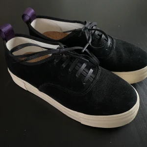 Svarta mockasneakers från Eytys - Snygga svarta sneakers från Eytys i mocka med vit platt sula och lila hälflik. Klassisk rund tå och svarta snören. Ikonisk korkinnersula med Eytys-logga. Perfekta för dig som gillar stilrena och trendiga skor.