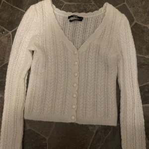 Vit stickad kofta från Bik Bok - Supersöt vit stickad kofta från Bik Bok med kabelstickat mönster och v-ringning. Koftan har långa ärmar, ribbade muddar och knappar framtill. Perfekt att slänga över en topp för en mysig look.