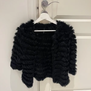 Svart pälsjacka med kort ärm - Säljer en svart pälsjacka från H&M med korta ärmar och fluffig, mjuk känsla. Jackan har en öppen front och ett coolt randigt pälsmönster som ger en lyxig vibe. Perfekt för dig som vill sticka ut och hålla dig varm med stil.