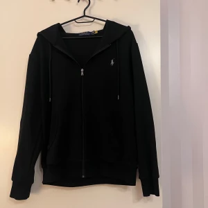Ralph Lauren zip up  - Svart zip up från Polo Ralph Lauren med dragkedja framtill, huva och snörning. Klassisk broderad logga på bröstet och två fickor framtill. Perfekt för en chill och stilren look.