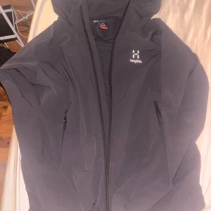 Svart vindjacka från Haglöfs - Säljer en svart vindjacka från Haglöfs med huva och hel dragkedja framtill. Jackan har två sidofickor med dragkedja och en stilren design med logga på bröstet. Perfekt för blåsiga dagar och outdoor-äventyr.