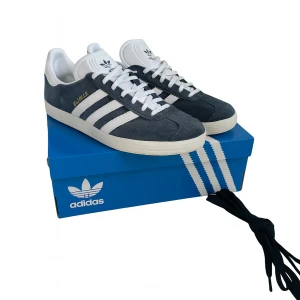 Adidas Gazelle - Säljer nu snygga Adidas Gazelle sneakers i blå mocka med vita detaljer och snörning!🤩🙌🏼!Skorna har den klassiska tre-randiga designen och kommer med extra skosnören. Perfekta för en stilren look. Box samt extra snören medföljer! Se bild 3 med de andra snörena! Byten intressant! kom med bud!🍾🍾