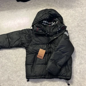 Svart dunväst från Burberry XL/L - Svart dunväst från Burberry med avtagbara ärmar, huva och klassiskt rutigt foder. Västen har dragkedja, tryckknappar och två fickor framtill. Stor logotyp på axeln och snygga detaljer. Perfekt för lager-på-lager-stil under kyliga dagar.