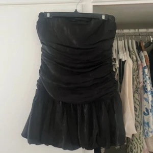 Svart axelbandslös kortklänning  - Supersnygg svart axelbandslös kortklänning från MNG. Klänningen har en tight passform upptill, draperad midja och volangig kjol. Använd en gång så den är precis som ny! Storlek XS men passar även en S🤗köpt för 799kr 