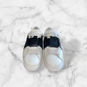 Valentino Open sneakers vit/svart - Snygga Valentino Open sneakers i vitt skinn med bred svart läderdetalj över mitten. Klassisk rund tå, vita snören och diskreta nitar baktill. Perfekta för dig som gillar stilrena och exklusiva sneakers med en twist. Verkligen stiliga och i riktigt bra skick! Hör av er för fler bilder eller frågor!
