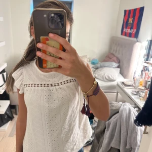 Vit blus med spets och volangärm - Säljer en söt vit blus i bomull med broderade detaljer och hålmönster. Blusen har volangärmar och en fin spetsrad över bröstet. Perfekt för dig som gillar romantisk och feminin stil. Rund halsringning och luftig passform.