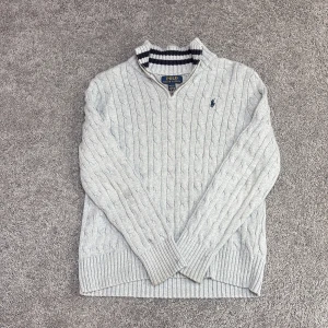 Grå kabelstickad tröja Polo Ralph Lauren - Kabelstickad grå tröja från Polo Ralph Lauren med halv dragkedja och marinblå logga på bröstet. Tröjan har ribbade muddar, lång ärm och en krage med marinblå/vita ränder på insidan. Perfekt för lager-på-lager-stil.