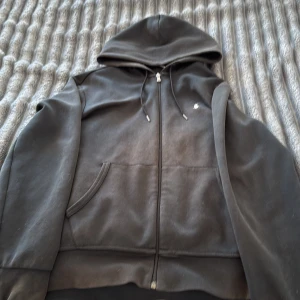 Svart hoodie från Polo Ralph Lauren - Svart hoodie från Polo Ralph Lauren med dragkedja och huva. Klassisk design med liten broderad logga på bröstet, känguruficka och snörning i huvan. Tillverkad i mjuk bomullsmix, perfekt för chill dagar eller när du vill ha en clean look.