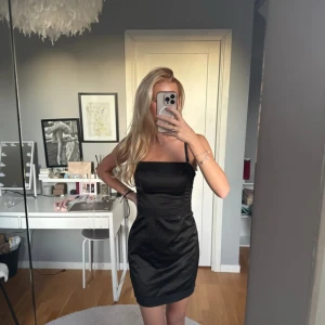 Svart kort klänning med spetsdetaljer  - Säljer en svart kort klänning med smala axelband och fina spetsdetaljer upptill, nedtill och i midjan. Banden är avtagbara och kan därför bli en bandeau klänning. Klänningen har en figurnära passform och är tillverkad i ett glansigt material som ger en lyxig känsla. Perfekt för dig som vill ha en stilren och elegant look.