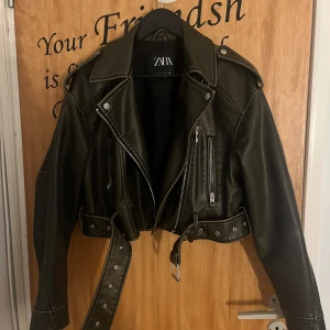  Croppad skinnjacka från Zara - Snygg croppad skinnjacka från Zara med bred krage, silverfärgade dragkedjor och detaljer samt bälte i midjan. Jackan har långa ärmar och cool bikerstil som ger en edgy vibe till din outfit.