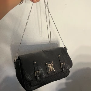Svart axelväska från Guess i skinn - Snygg svart axelväska från Guess med reptilmönstrad yta i skinn. Väskan har silverfärgade detaljer, dubbla spännen framtill och en kedja som axelrem. På framsidan sitter Guess-loggan i metall. Perfekt storlek för det viktigaste och ger en cool vibe.