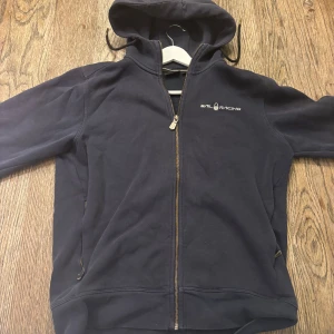 Sail Racing hoodie - Svart hoodie från Sail Racing med dragkedja framtill och broderad logga på bröstet. Tröjan har huva med snörning och två fickor med dragkedja. Perfekt för dig som gillar en sportig och stilren look.