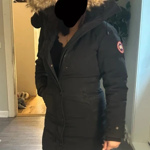 Canada Goose jacka - Jackan har lång passform, dragkedja och knappar framtill samt två fickor. Perfekt för kalla dagar och har den klassiska loggan på ärmen. Pälsen går att ta av och man kan justera jackan så att den passar ens kropp. Jackan är använd men har inte mer än hålen på ärmarna och ett hål på insidan av ena fickan!