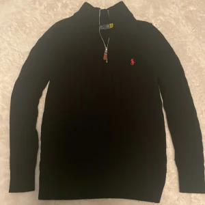 Svart half zip tröja Polo Ralph Lauren - Svart långärmad tröja från Polo Ralph Lauren med röd broderad logga på bröstet. Tröjan har en snygg half zip-dragkedja med läderdetalj och hög krage. Perfekt för dig som gillar klassisk och stilren design.
