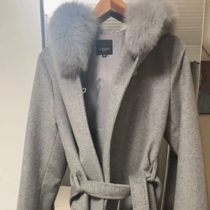 Grå kappa med päls från Hollies - Snygg grå kappa från Hollies med stor fluffig päls runt huvan och knytskärp i midjan. Klassisk och stilren modell i ullmix, perfekt för kyliga dagar. Långärmad och med en mjuk känsla i materialet.
