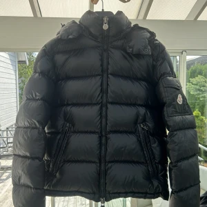 Svart Moncler Maya - Säljer en svart pufferjacka från Moncler med glansig finish, vadderad design och avtagbar huva. Jackan har dragkedja framtill, två snedställda fickor med dragkedja och Moncler-logga på ärmen. Perfekt för kalla dagar och riktigt snygg streetstil.