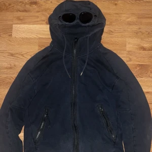 Svart hoodie med goggles från C.P. Company - Svart hoodiejacka från C.P. Company med ikoniska goggles i huvan. Jackan har dragkedja framtill, två snedställda fickor med dragkedja och justerbar huva med snörning. Tillverkad i mjuk bomull för en skön och avslappnad look. Size XS, 8/10! Fler frågor i pm/Neo💫