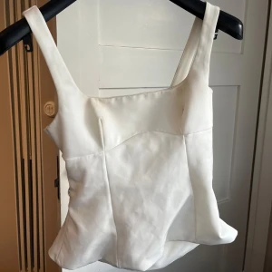 Vit korsett-topp med knyt i ryggen - Säljer en vit korsett-topp med breda axelband och snyggt formad siluett. Toppen har ett coolt knyt i ryggen som ger en öppen look och extra detalj. Materialet är slätt och har en lätt glans, vilket gör den extra fräsch. Perfekt för dig som gillar stilrena och trendiga plagg.