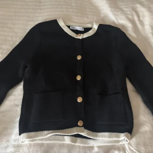 Svart kofta från Zara med guldfärgade knappar - Svart kofta från Zara med kontrasterande vit kant vid hals och nederkant. Koftan har guldfärgade knappar framtill och två fickor. Stickad i mjukt material och har långärmad passform. Perfekt för att addera lite stil till din outfit.