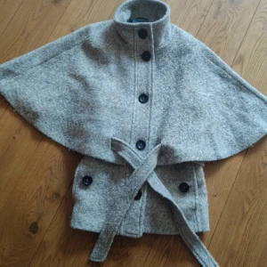 Grå cape med bälte och knappar - Snygg grå cape-kappa i ullmix med hög krage, stora svarta knappar framtill och tillhörande bälte i midjan. Är perfekt för höst och vinter. SÅÅ himla snygg men har en lika dan i en mörkare färg! Båda ligger ute så behåller den som inte säljs!🍁 kolla andra annonsen i min profil för att se bild på passformen! OBS inte exakt samma jacka men sitter exakt lika! Storlek M men passar mig som brukar ha XS och S, passar även M!